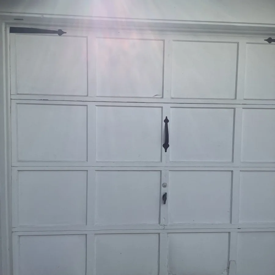 A white carriage-style garage door with square recessed panels, decorative strap hinges and a spear handle, photographed close up with sunlight flaring across the top corner.