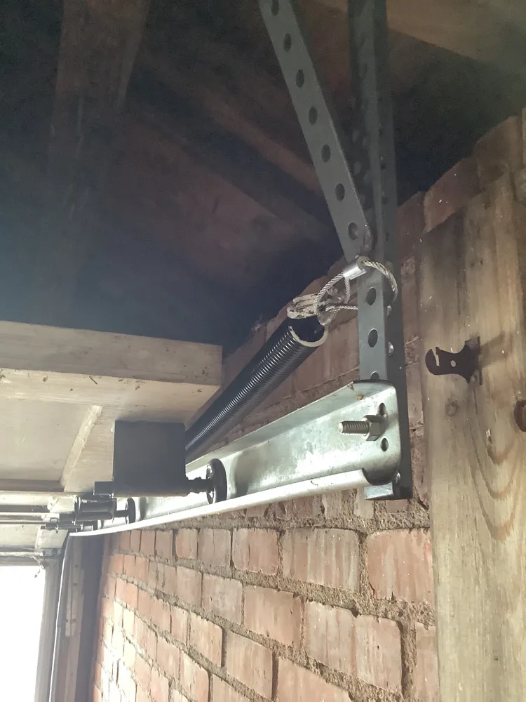 An extension spring and its cable strung along an angle-iron track above a garage opening, bolted through a brick wall beside the timber jamb.