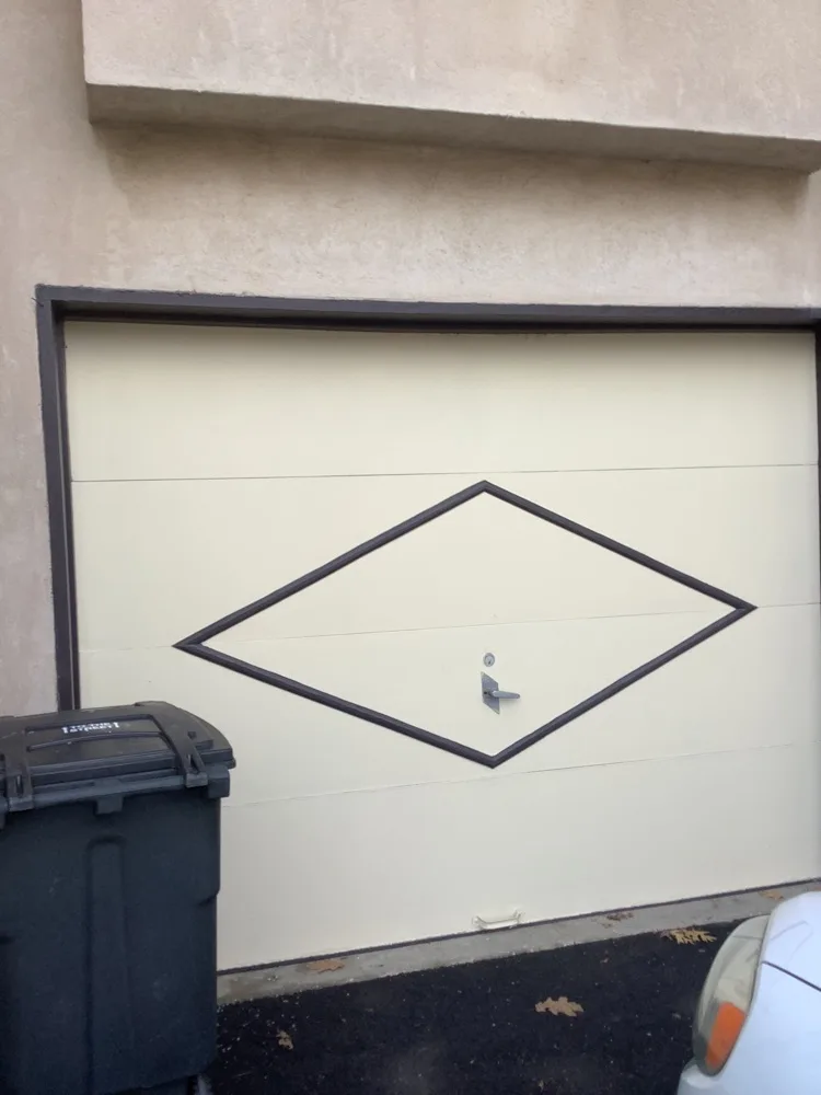 A cream flush-panel garage door with a black diamond overlay and a centre handle, set into a stucco wall with a wheelie bin standing at the left edge of the opening.