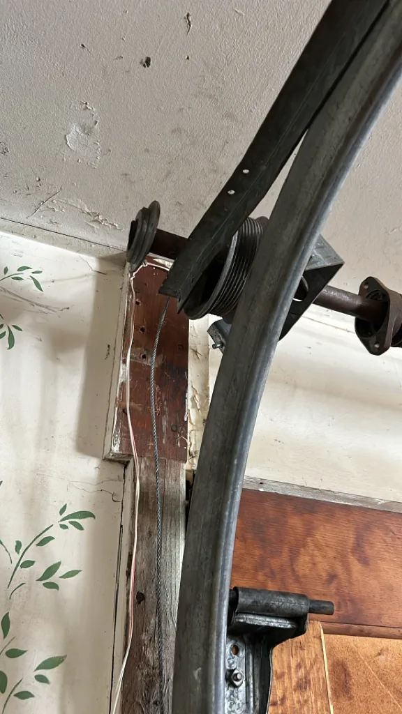 A lift cable running down from a grooved drum and end bearing at the top corner of a garage door, alongside the curved section of track where the rollers turn.