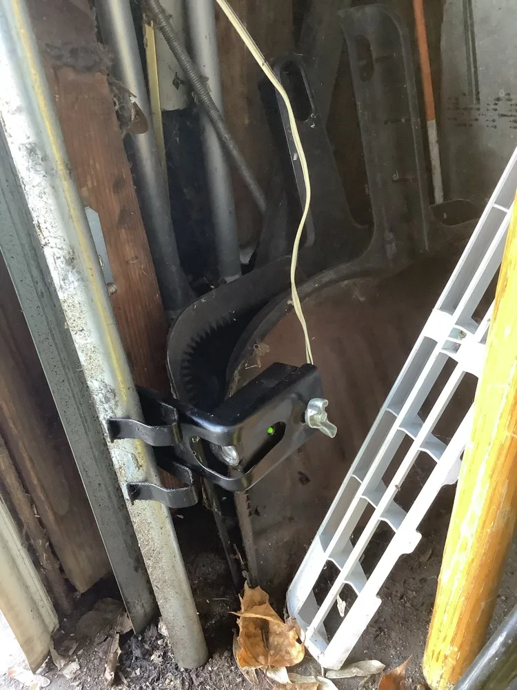 A photo-eye safety sensor showing a green indicator light on its bracket at the foot of a garage door track, with dead leaves and grit collected around the rail.