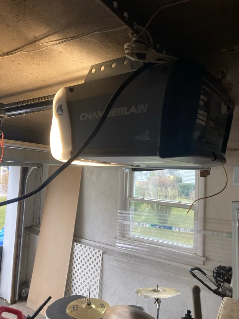 A chain-drive opener mounted low under a garage ceiling, its light on, with a side window looking out to a green yard behind it.