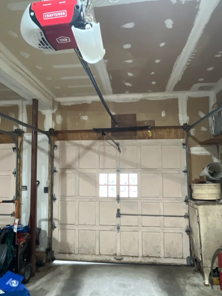 A garage interior with a red and white opener bolted to the ceiling, its rail running down to a closed panelled door with a single small window.