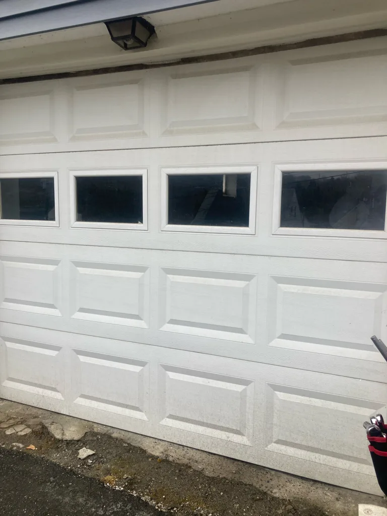 A white raised-panel garage door photographed square on, with four rectangular window lites across the upper section and a carriage lamp above the head.