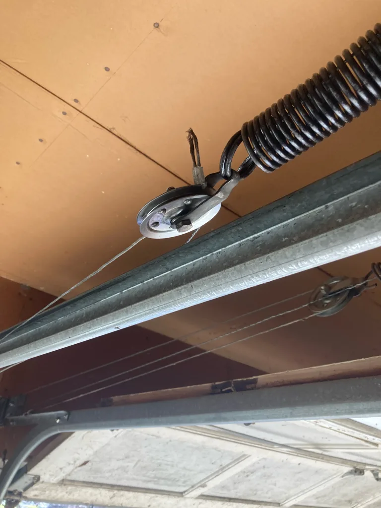An extension spring hooked to a pulley at the ceiling above a garage door, its lift cable running down over the sheave to a second pulley further along the track.