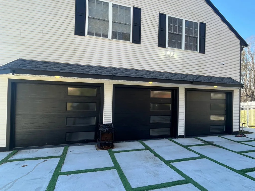 Three matte-black flush-panel garage doors with vertical rows of frosted glass lites, set into a cream clapboard house under a clear blue sky, with a paver-and-turf driveway running up to them.